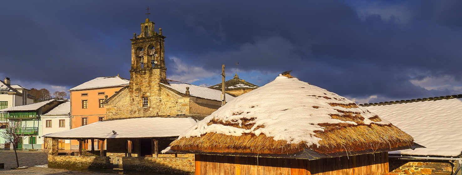 Vista de Samartín, capital del municipio de San Martín de Oscos, en un día invernal. Vemos una iglesia de piedra, con su torre campanario, dominando la plaza principal del pueblo, y junto a ella, un tradicional hórreo de madera, con los tejados cubiertos de un manto blanco de nieve, que añade un toque rústico y pintoresco.