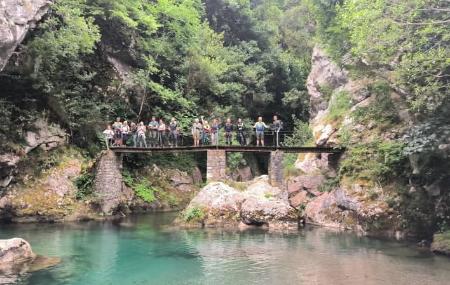 A group of people stand on a narrow wooden bridge supported by stone pillars, set between rocky walls and dense vegetation. A clear pool of water flows under the bridge in a gorge-like setting.