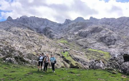 Three people equipped for hiking stand in an area of alpine grassland, surrounded by a landscape of limestone rock and jagged slopes rising towards a line of mountain ridges. In the background is a small hut built into the terrain, under a partly cloudy sky.