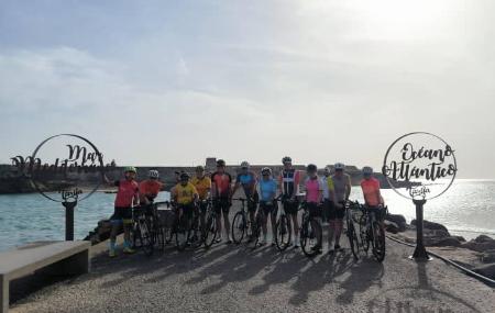 A group of cyclists pose with their bicycles on a coastal breakwater, flanked by two circular structures marked 