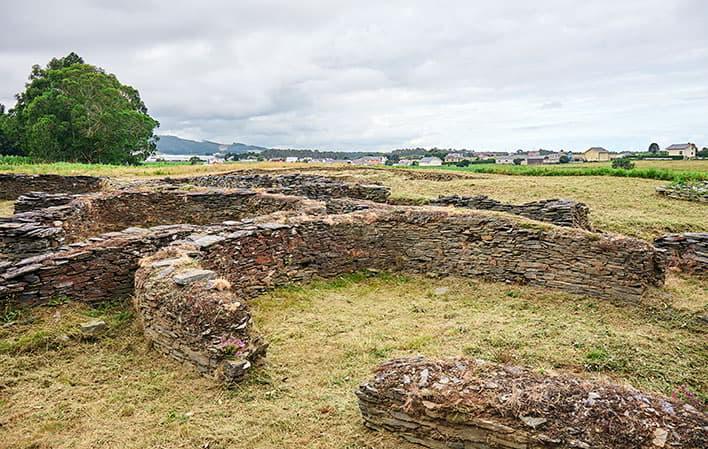 Restos arqueológicos del Castro de Mohías con muros bajos de piedra formando estructuras circulares y rectilíneas sobre un terreno de hierba segada, rodeados de campos de cultivo y algunas edificaciones al fondo bajo un cielo nublado.