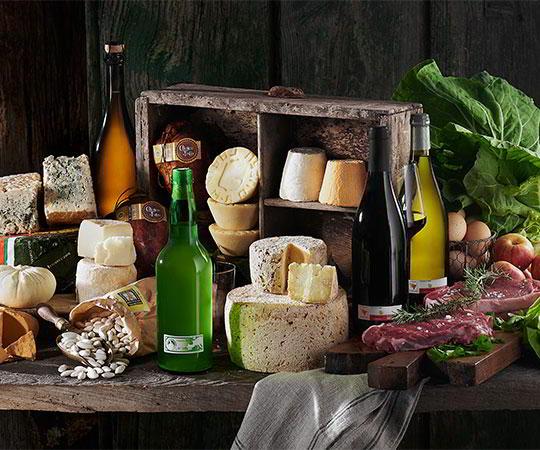 Photo of a still life arrangement featuring various Asturian products. Different types of Asturian cheeses, meat, fabas (white beans), cured meats, cider, and Cangas wine