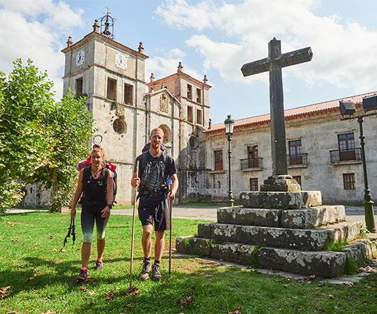 Photo d'un couple de pèlerins en premier plan, sur le Chemin de Saint-Jacques Primitif, avec le Monastère de San Salvador dans la commune de Salas
