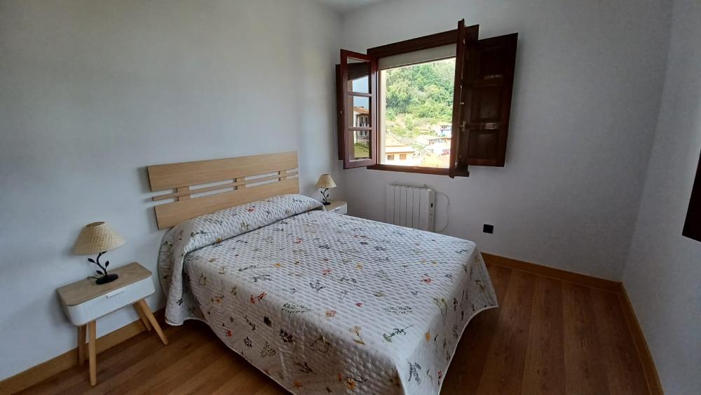 Dormitorio luminoso con cama de matrimonio, dos mesitas de noche y ventana abierta con vistas al exterior.