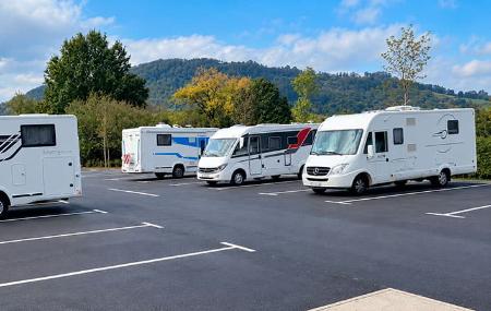 Varias autocaravanas estacionadas en las plazas habilitadas del área de Lugones, rodeada de árboles y montañas.