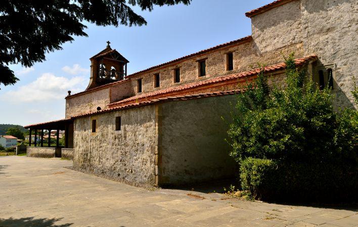 A igreja pré-românica de Santiago de Gobiendes num dia de sol, com uma árvore em primeiro plano e a estrada de acesso à igreja.